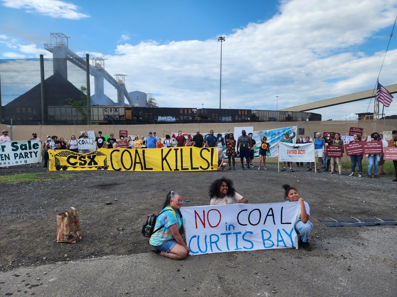 Residents of South Baltimore, local activists and supporters from around the city gather in front of the CSX Curtis Bay Pier in Curtis Bay, Baltimore—with a CSX locomotive and giant coal piles visible in the background—and pose for a picture with signs reading, “No Coal in Curtis Bay,” “CSX COAL KILLS,” etc. Photo by Maximillian Alvarez