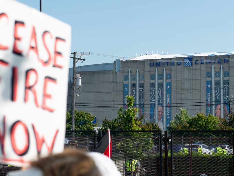 Image of a protest sign saying "CEASE FIRE NOW" in the foreground with the United Center, the site of the 2024 Democratic National Convention, visible in the background. Photo taken by Steel Brooks in Chicago, IL, on Monday, Aug. 19