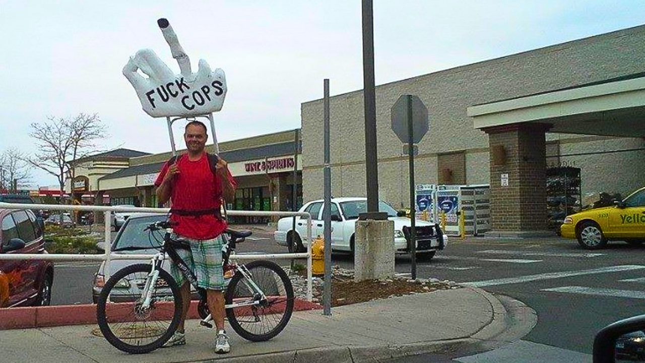 Photo of Eric Brandt exercising his First Amendment rights in Denver, Colorado, posing on a bicycle holding a giant hand over his head, which is pointing a middle finger and has "FUCK COPS" written on it. Photo courtesy of Eric Brandt.