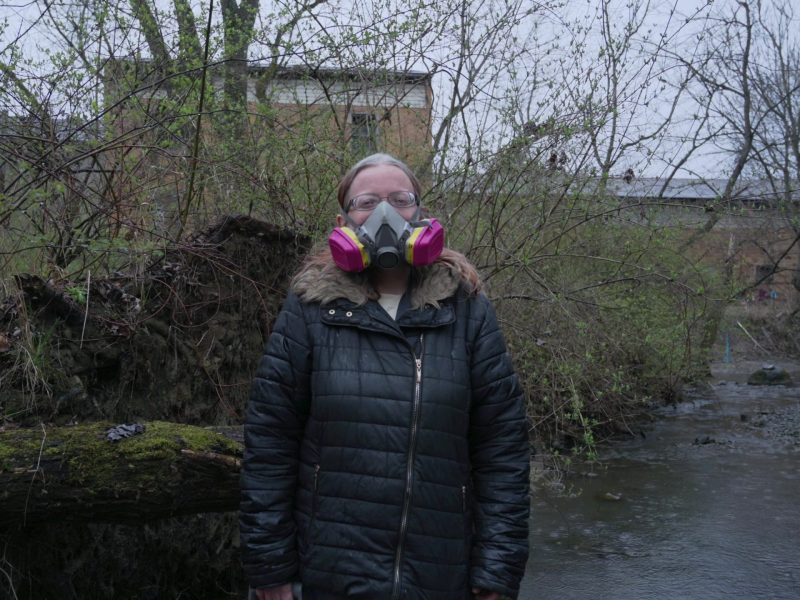 Still image from TRNN's documentary report "Trainwreck in 'Trump Country,'" featuring Christina Siceloff, a resident of Beaver County, PA, standing in a creek in East Palestine, OH, with a pink gas mask on her face. Image courtesy of Mike Balonek.