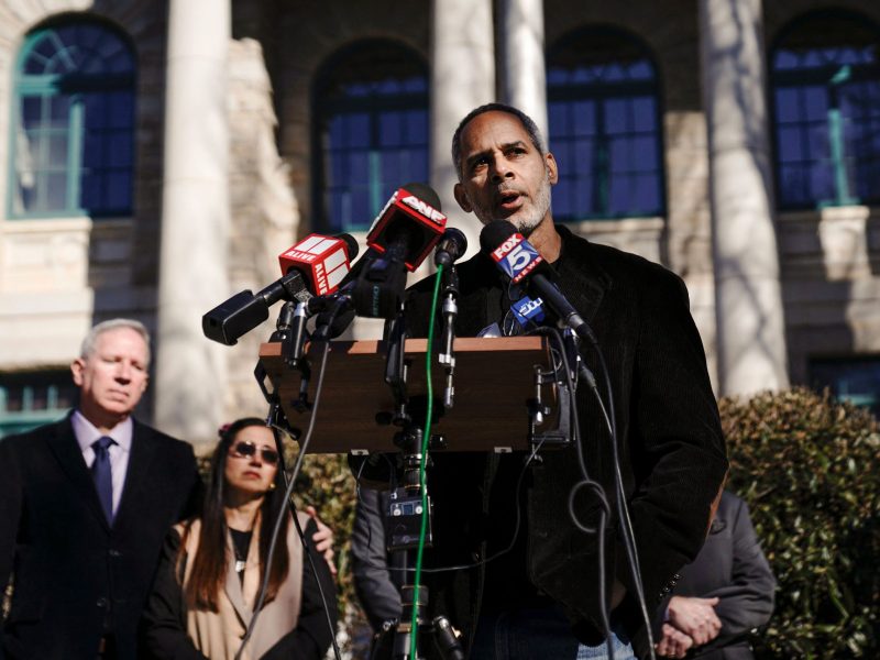 Kamau Franklin stands at a podium outdoors and speaks into a group of microphones. Behind him are Manuel Teran's parents, dressed in formal wear.