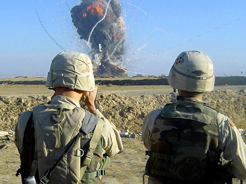 Two helmeted US soldiers seen from the back, looking into binoculars at a small mushroom cloud in the distance.