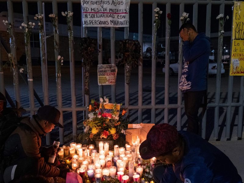 Migrants huddle around a makeshift altar of lit candles in the dark in front of a fence.