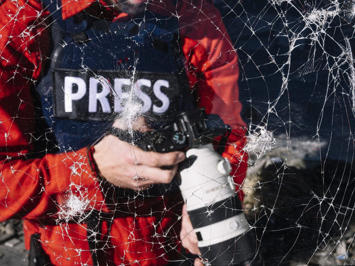 A war journalist wearing a vest labeled "Press" and holding a digital camera.