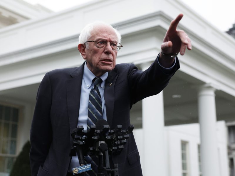 Senator Sanders speaks in front of the white columns of the White House's West Wing. He is wearing a dark suit and has one finger raised in the air.