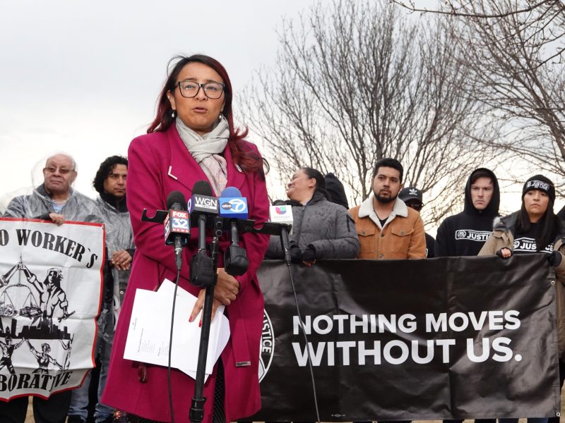 A woman in a red coat speaks into a microphone. Behind her are activists carrying two banners. One is a union banner, the other reads "Nothing moves without us."