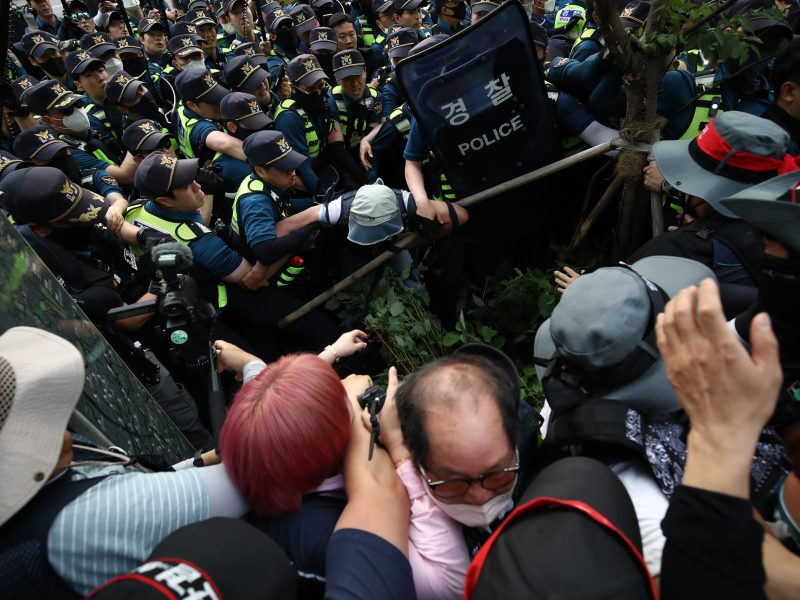 A wave of riot-shield wielding police officers descends upon a group of trade unionists, who have their arms raised in self defense.