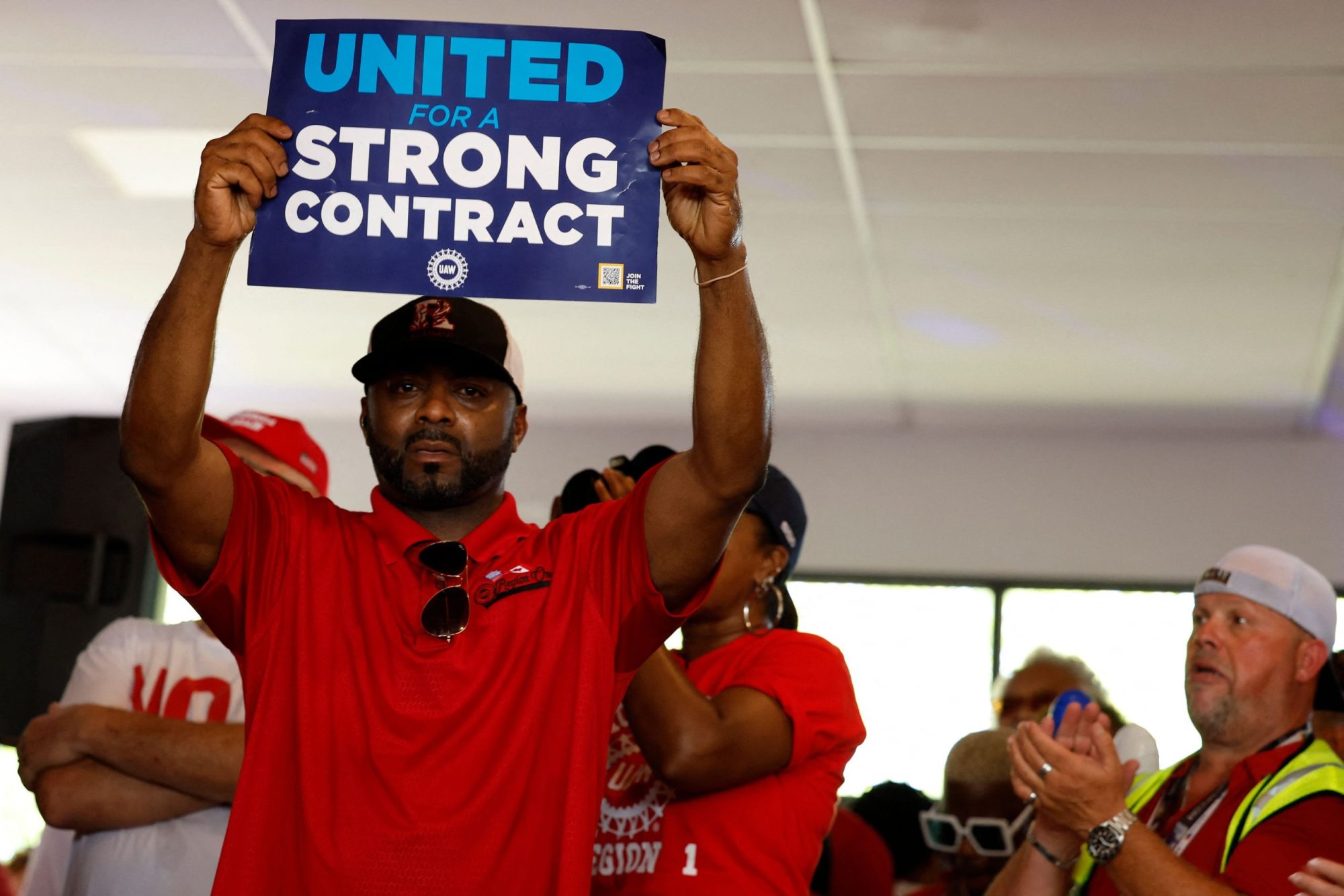 A worker in a red shirt holds up a sign that reads "United for a Strong Contract"