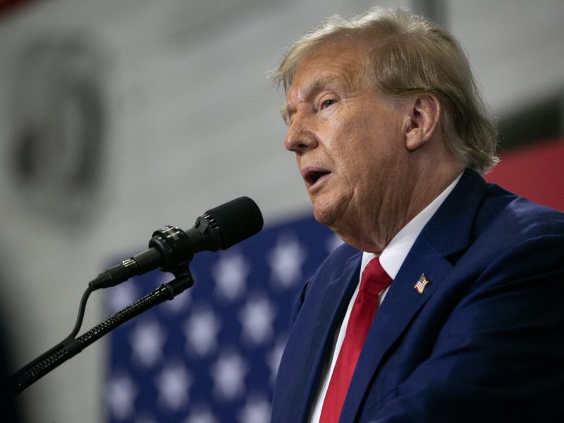 Donald Trump wearing a dark suit and red tie, speaking into a microphone in an indoor setting with the American flag in the background