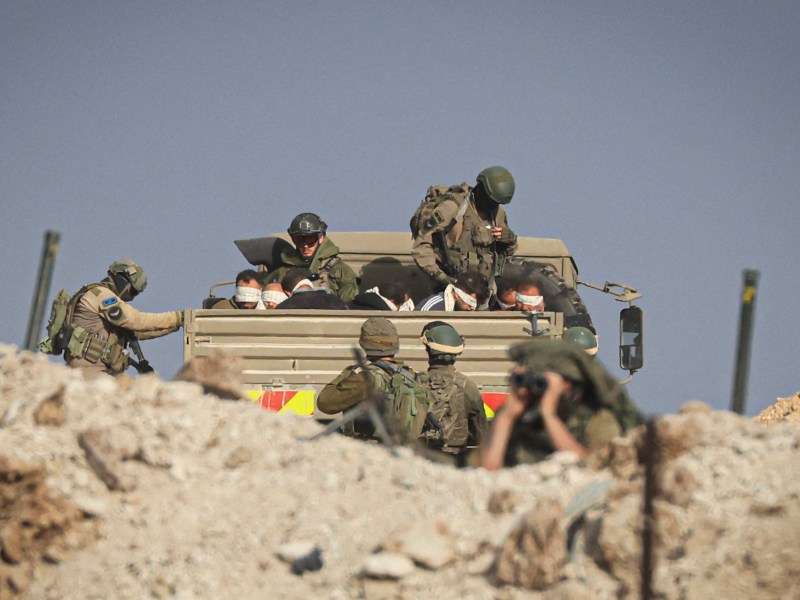 Israeli soldiers detain blindfolded Palestinian men in a military truck while watching Palestinians (not pictured) fleeing the fighting in war-torn Gaza walk by on a road in the Zeitoun district of the southern part of the Gaza Strip on November 19, 2023. Photo by MAHMUD HAMS/AFP via Getty Images