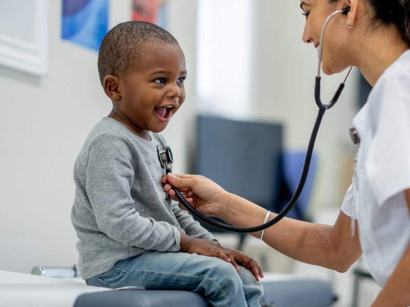 A young boy sits up on an exam table as a pediatrician preforms a check-up on him. Photo via Getty Images