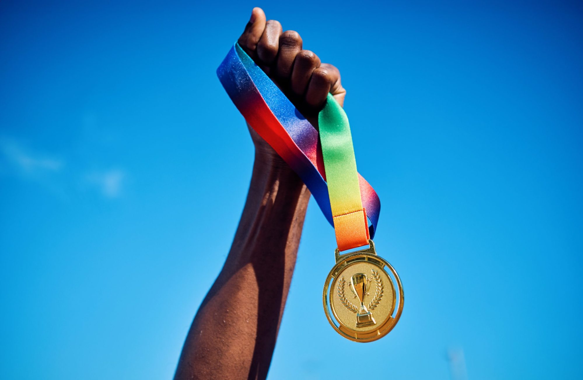 Raised hand holding a gold medal against blue sky. Photo via Getty Images