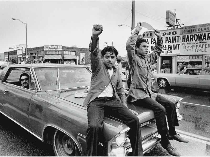 Two young Chicano men ride on the hood of a car and raise their fist during a National Chicano Moratorium Committee march in opposition to the war in Vietnam, Los Angeles, California, February 28, 1970. Photo by David Fenton/Getty Images