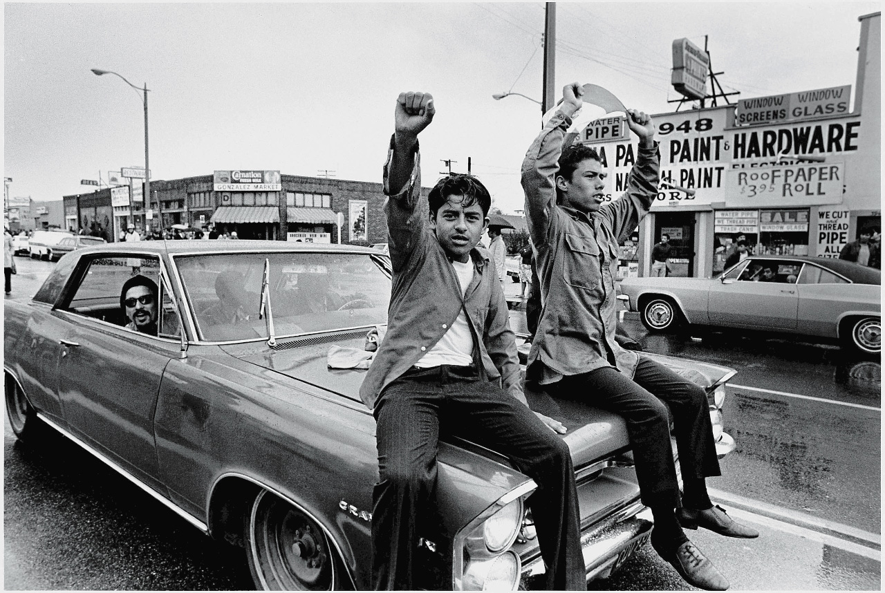 Two young Chicano men ride on the hood of a car and raise their fist during a National Chicano Moratorium Committee march in opposition to the war in Vietnam, Los Angeles, California, February 28, 1970. Photo by David Fenton/Getty Images