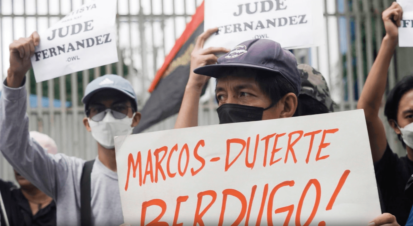 A man wearing a face mask and a hat holds up a sign that says "Marcos-Duterte Berdigo!" In the background two protestors wearing face masks, sunglasses, and caps hold up signs that read "Jude Fernandez"